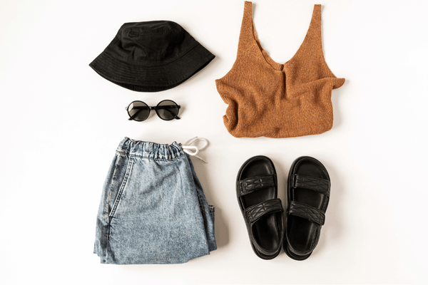 A photo in a white background showing a minimalistic outfit if a black hat, black glasses, black sandals, short jeans, and a brown sweater